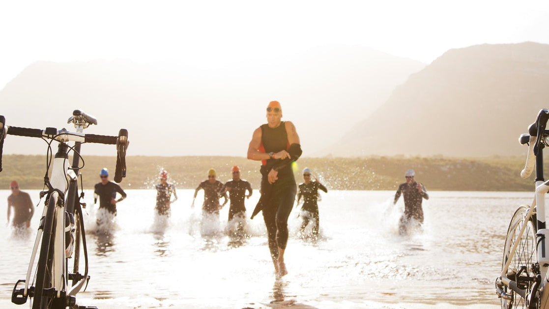 A group of male and female triathletes in swimsuits emerging from the water and running towards two bikes on a beach.